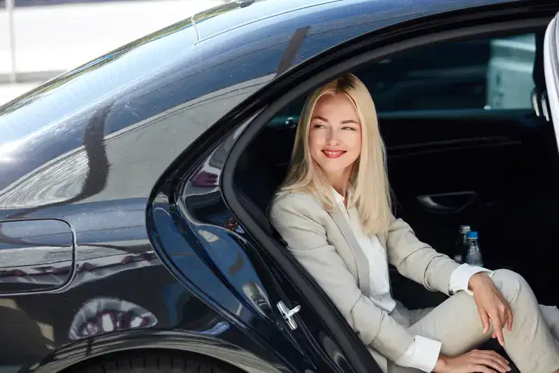 Professional woman stepping out of the rear seat of a black luxury car, smiling while wearing a light-colored business suit, with a water bottle visible inside the vehicle. Symbolizing Monterrey Luxury Transportation.